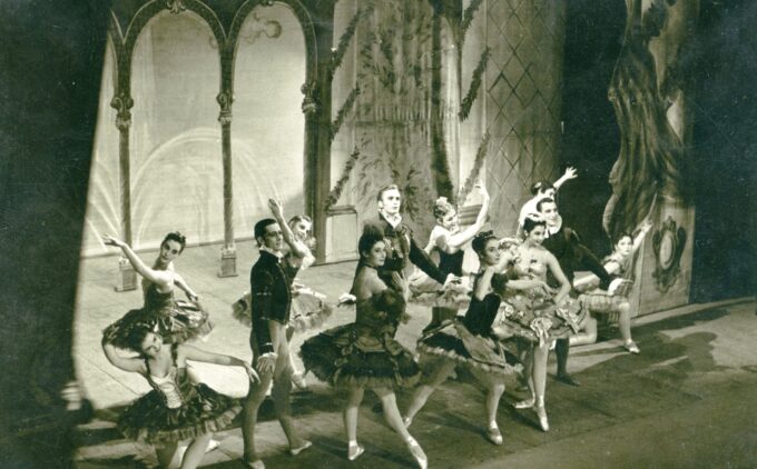A group of ballet dancers performs on stage in intricate costumes, with a detailed backdrop and theatrical lighting.