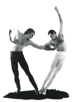 Two male ballet dancers perform a dynamic pose, with one leaning forward and the other arching backward, on a plain background.