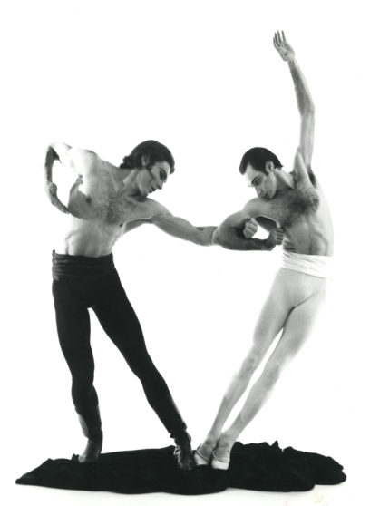Two male ballet dancers perform a dynamic pose, with one leaning forward and the other arching backward, on a plain background.