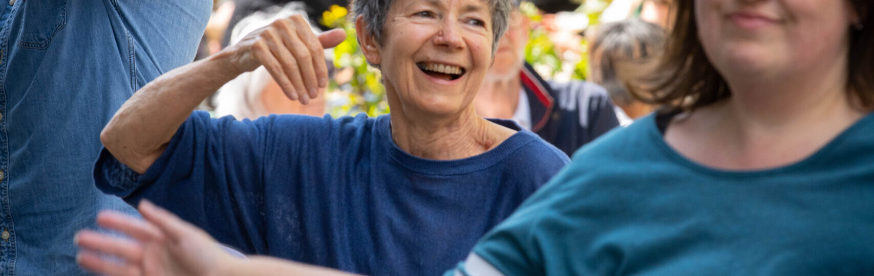 A group of people outdoors, smiling and dancing together in casual clothing. The background shows trees and a small crowd.