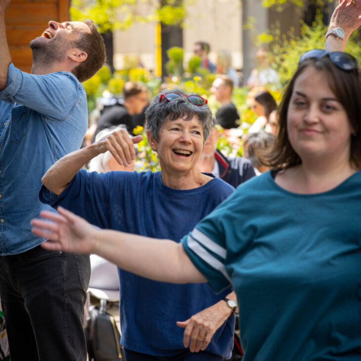 A group of people outdoors, smiling and dancing together in casual clothing. The background shows trees and a small crowd.