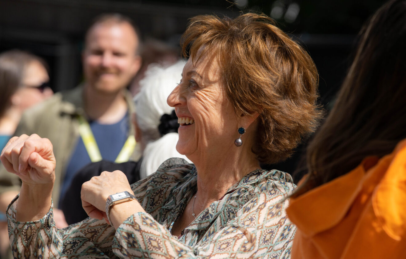A woman with short brown hair wearing a patterned blouse smiles and dances among a group of people in an outdoor setting.