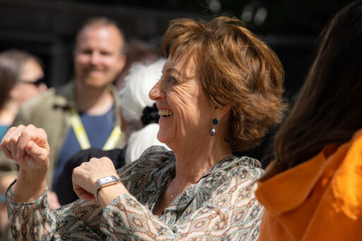 A woman with short brown hair wearing a patterned blouse smiles and dances among a group of people in an outdoor setting.