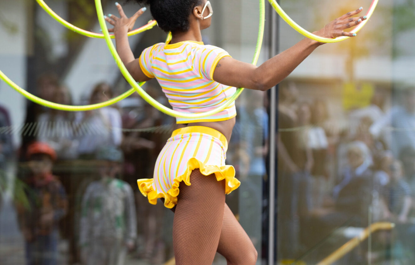 A performer in a yellow-striped outfit skillfully manipulates multiple hula hoops in an outdoor setting.