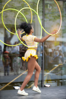 A performer in a yellow-striped outfit skillfully manipulates multiple hula hoops in an outdoor setting.