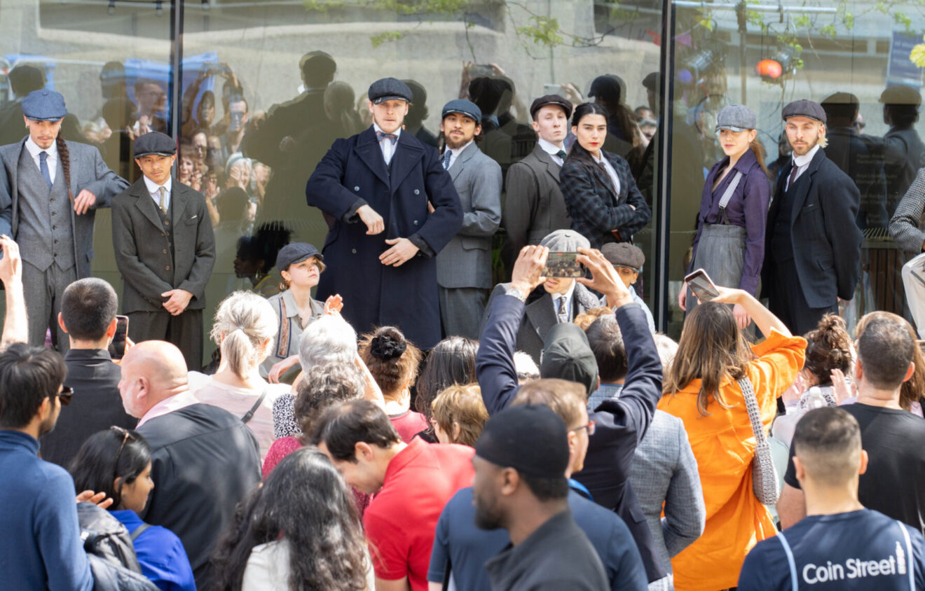 A group of people dressed in early 20th-century attire stands in front of a glass building, posing for a crowd. The crowd includes people taking photos and spectators looking at the group.
