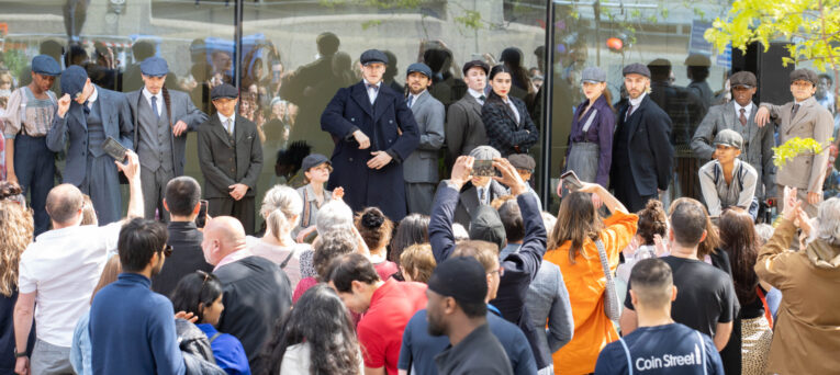A group of people dressed in early 20th-century attire stands in front of a glass building, posing for a crowd. The crowd includes people taking photos and spectators looking at the group.