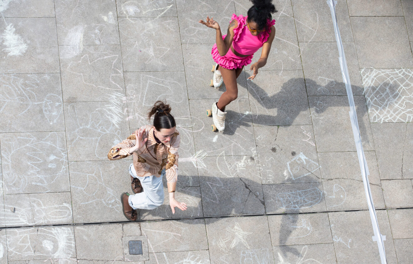 Two people dressed in bright outfits roller skate on a chalk-drawn pavement, viewed from above. The ground is decorated with various chalk drawings.