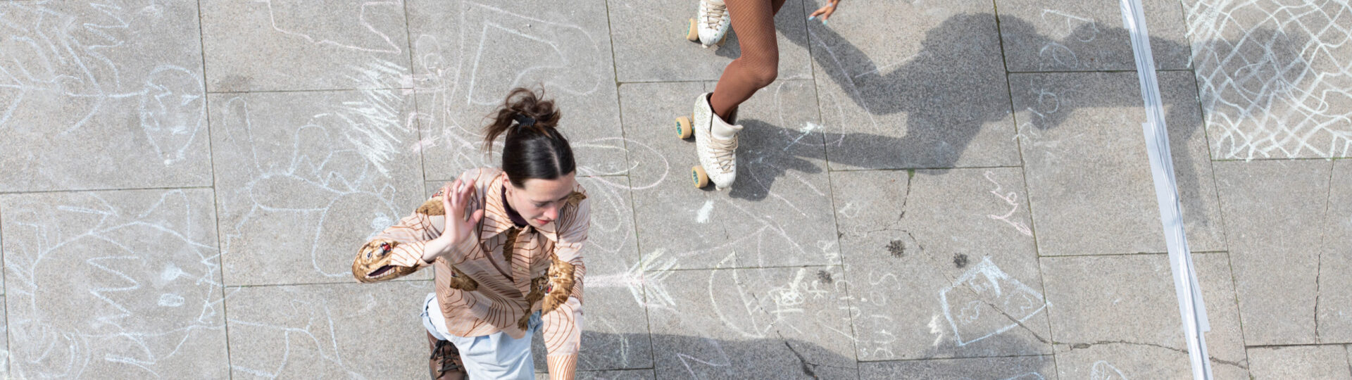 Two people dressed in bright outfits roller skate on a chalk-drawn pavement, viewed from above. The ground is decorated with various chalk drawings.
