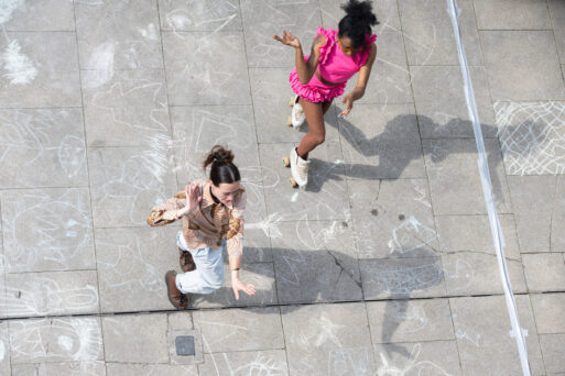 Two people dressed in bright outfits roller skate on a chalk-drawn pavement, viewed from above. The ground is decorated with various chalk drawings.