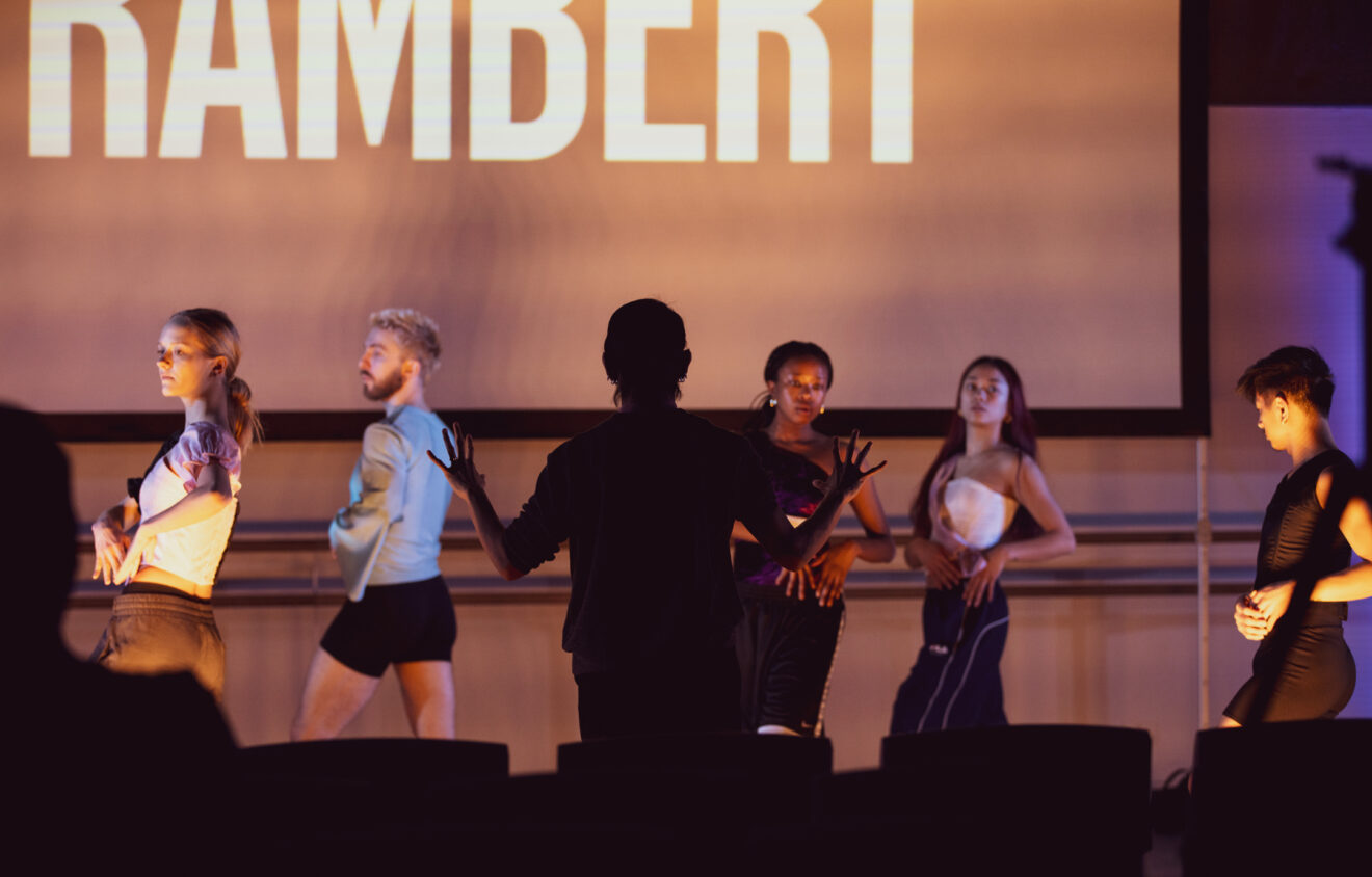 A group of dancers rehearses on stage with a person in front guiding them. The word "RAMBERT" is displayed on a screen behind them.