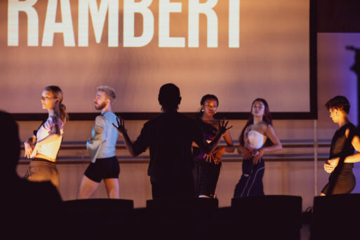 A group of dancers rehearses on stage with a person in front guiding them. The word "RAMBERT" is displayed on a screen behind them.