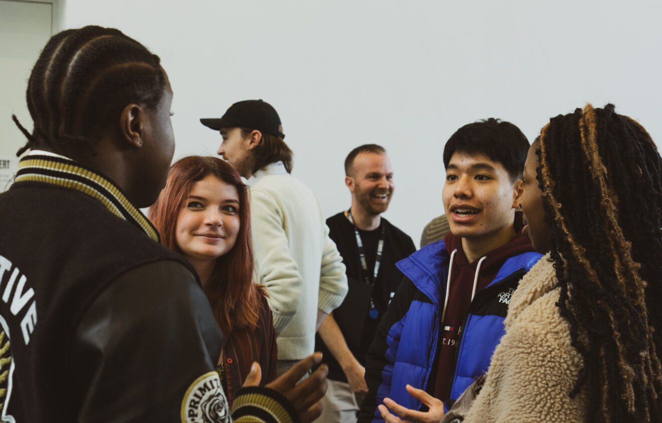 A group of people engaged in conversation indoors; one person wears a black and white jacket, another with red hair smiles, and a third person in a blue jacket speaks to another wearing a tan coat.