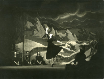 A dancer performs in the spotlight on a dark stage with a dramatic, mountainous backdrop. Three seated individuals in shadowed costumes observe from the sides.