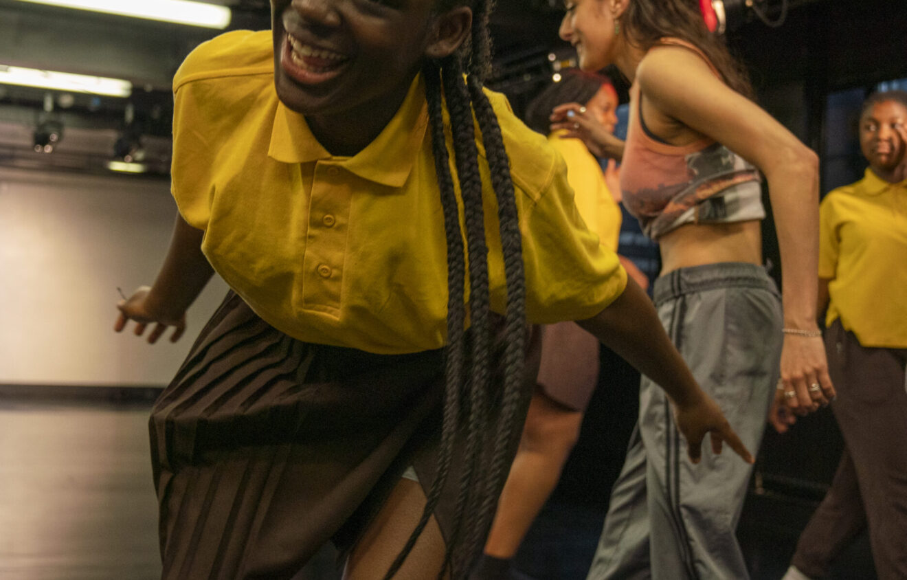 A person wearing a yellow shirt and brown skirt smiles and leans forward while dancing in a studio, with others dancing in the background.