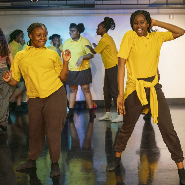 A group of people in yellow shirts and dark pants dance energetically on a stage with a polished floor.