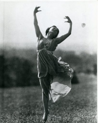 A dancer wearing a flowing dress performs a graceful leap outdoors on a grassy field.