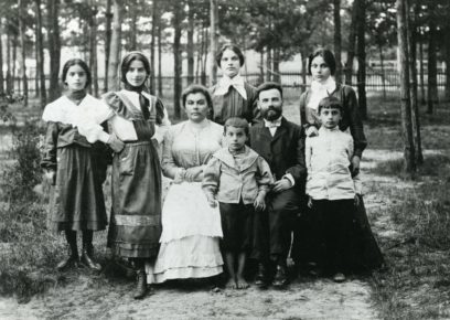 A black-and-white photo of a family with three adults and five children. They are posing outdoors in front of a forested area, with the adults seated and the children standing and sitting around them.