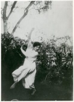 A woman in a white dress dances outdoors among trees and foliage.