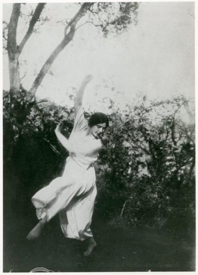 A woman in a white dress dances outdoors among trees and foliage.