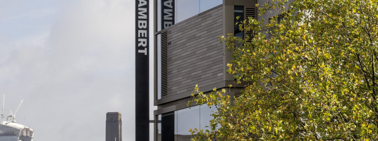 A modern building with "Rambert" signage, surrounded by cityscape and tree foliage in the foreground. The sky is partly cloudy.