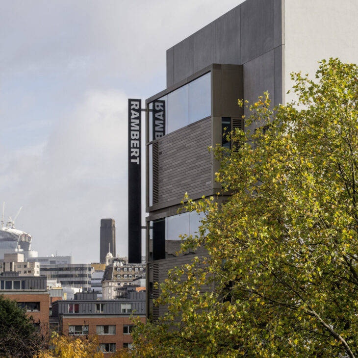 A modern building with "Rambert" signage, surrounded by cityscape and tree foliage in the foreground. The sky is partly cloudy.