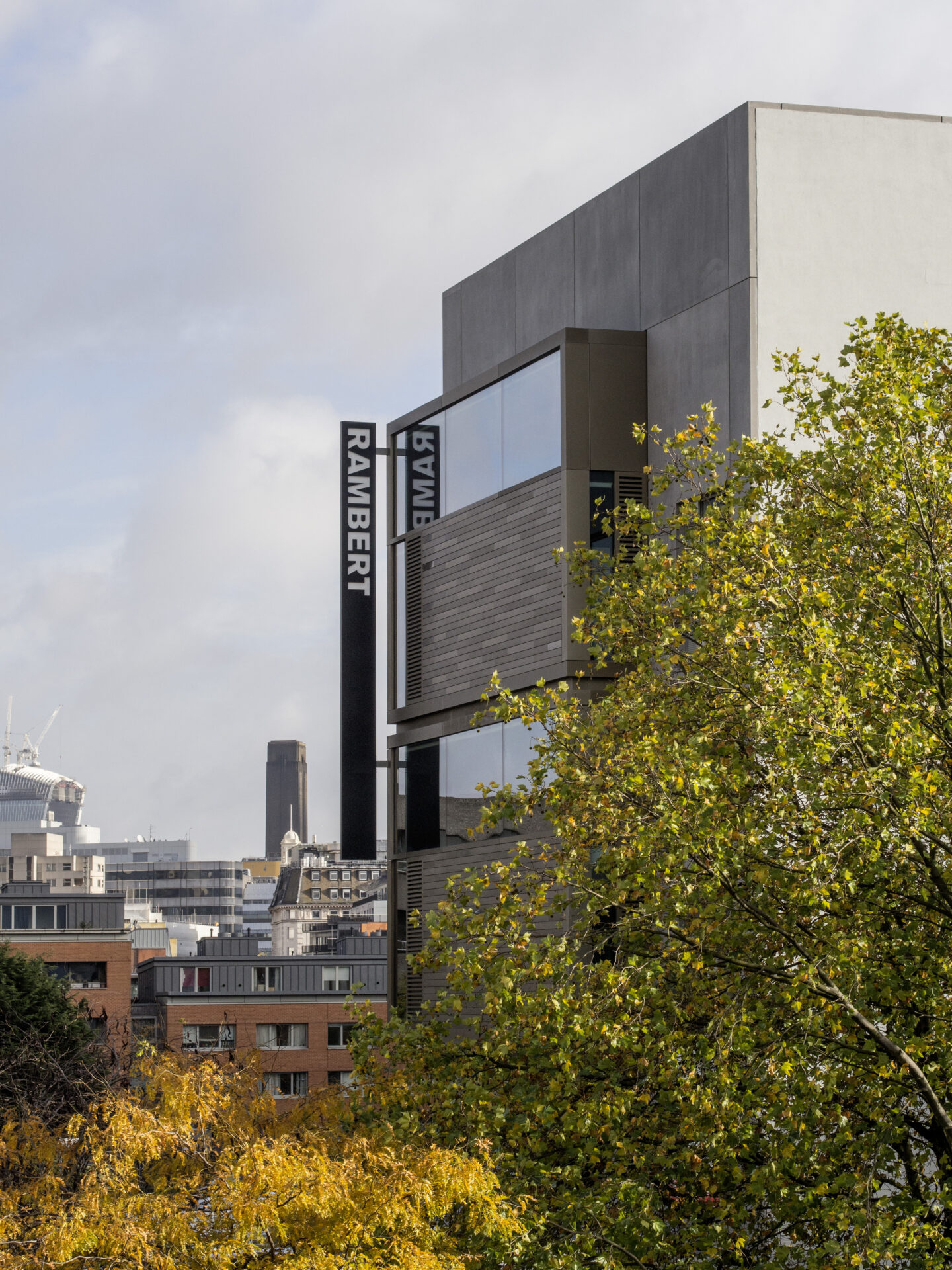 A modern building with "Rambert" signage, surrounded by cityscape and tree foliage in the foreground. The sky is partly cloudy.