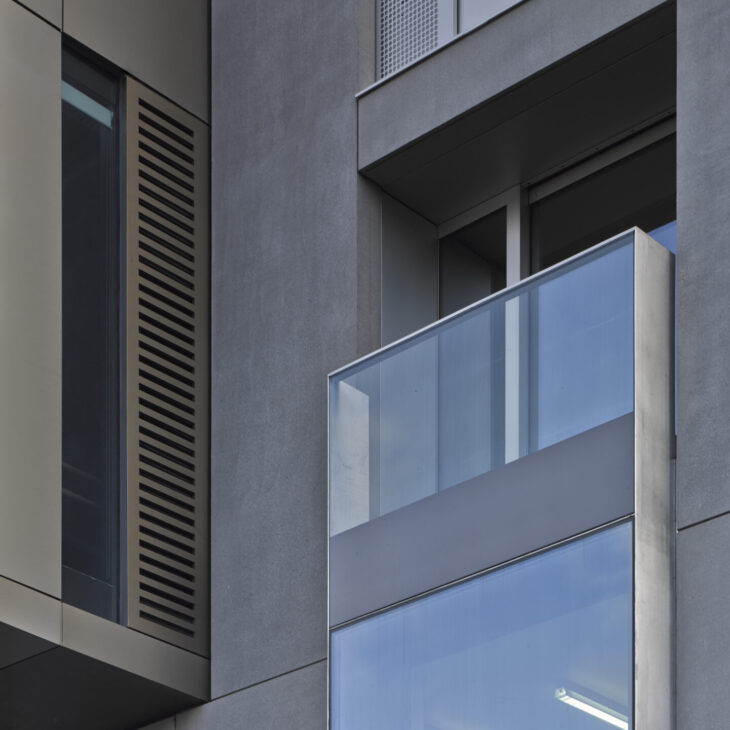 A modern building facade featuring a glass balcony, large window panels, and a vertical ventilation grille. The structure is composed of sleek, clean lines and a minimalist design.