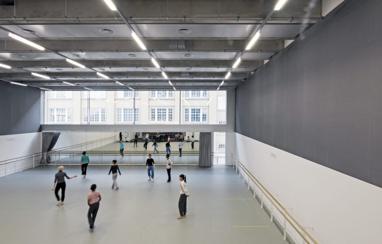 A spacious dance studio with high ceilings, large mirrors, and natural light from wall-length windows. Several people are practicing dance, dispersed throughout the room.