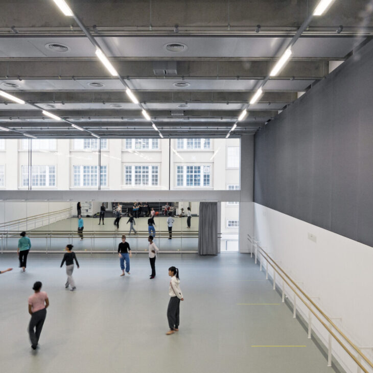 A spacious dance studio with high ceilings, large mirrors, and natural light from wall-length windows. Several people are practicing dance, dispersed throughout the room.
