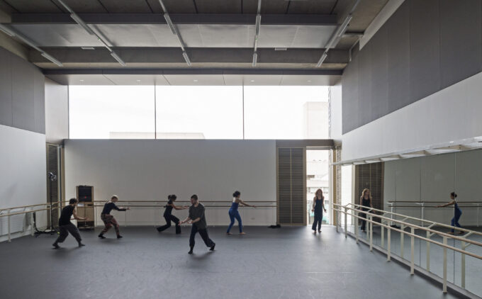 A group of people practice ballet movements in a spacious dance studio with mirrors and large windows.