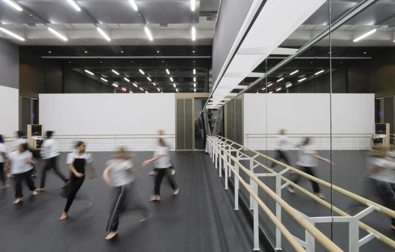 A group of dancers in motion during a rehearsal in a large, modern studio with floor-to-ceiling mirrors and ballet barres. The studio is well-lit with overhead lighting.