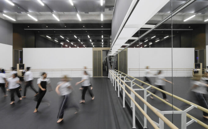 A group of dancers in motion during a rehearsal in a large, modern studio with floor-to-ceiling mirrors and ballet barres. The studio is well-lit with overhead lighting.