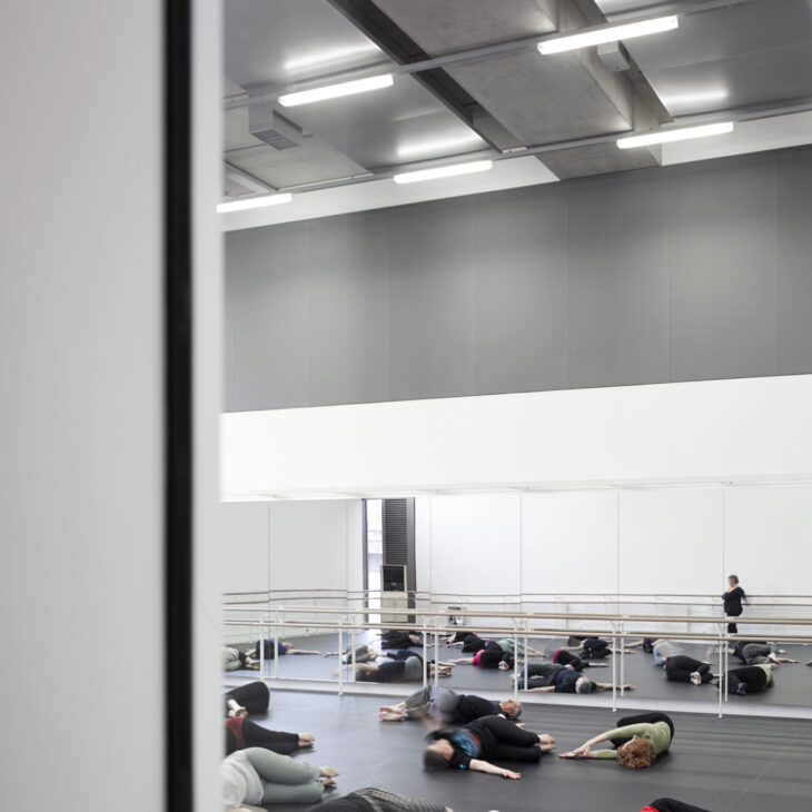A group of people lying on a dance studio floor, stretching, with a mirrored wall and barre visible, under bright overhead lighting.