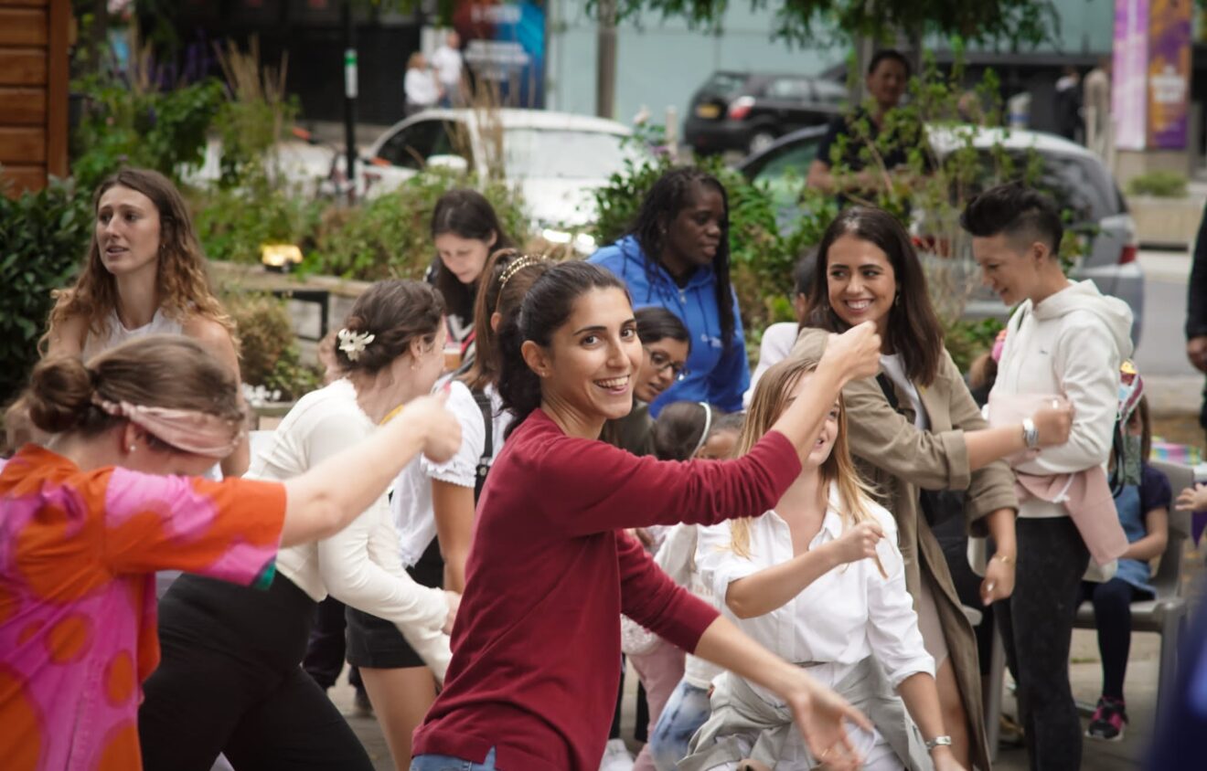 A group of people, including adults and children, are gathered outdoors, smiling and engaging in energetic dancing. Some are wearing colorful outfits. Trees and buildings are visible in the background.