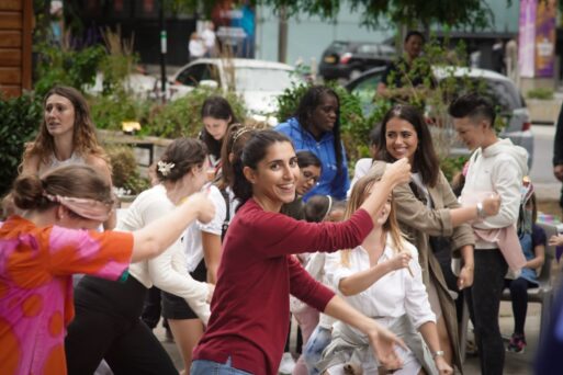 A group of people, including adults and children, are gathered outdoors, smiling and engaging in energetic dancing. Some are wearing colorful outfits. Trees and buildings are visible in the background.