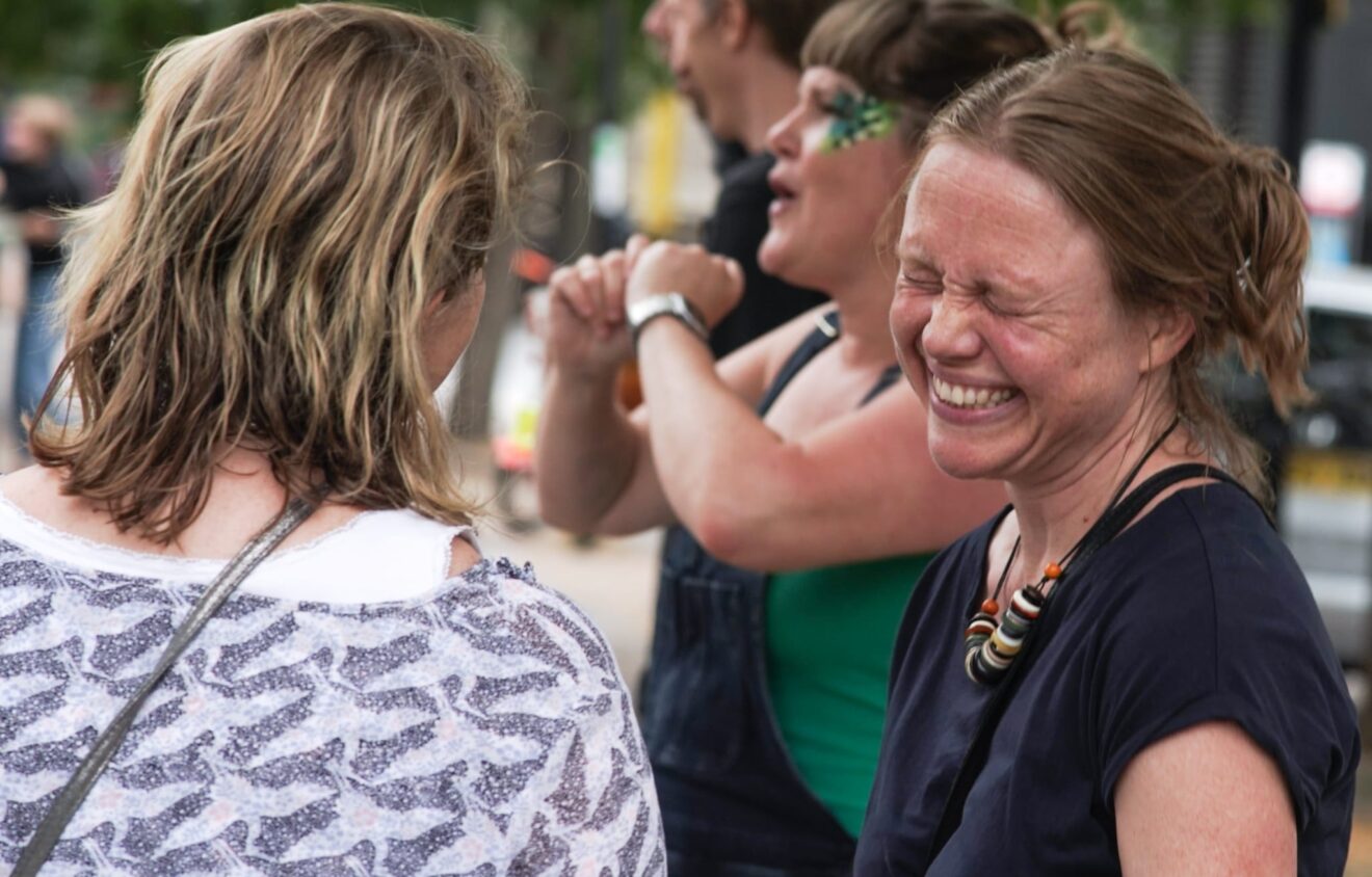 Four people are outdoors in a casual setting, with one woman smiling broadly and the others engaged in conversation.