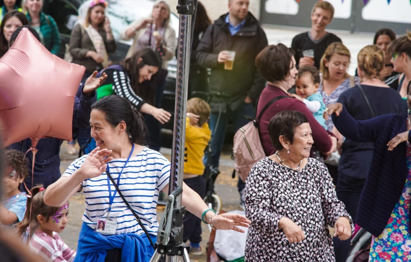 A diverse group of people, including children and adults, enjoy a lively outdoor gathering with balloons and festive decorations. Two women in the foreground appear to be dancing.