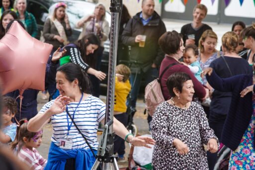 A diverse group of people, including children and adults, enjoy a lively outdoor gathering with balloons and festive decorations. Two women in the foreground appear to be dancing.