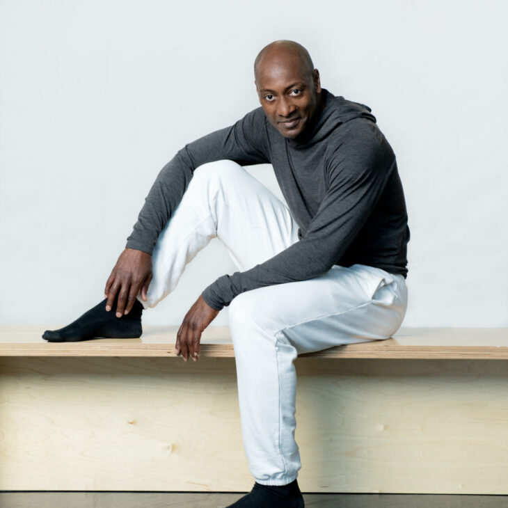 A man sits on a bench, wearing a long-sleeved gray shirt, white pants, and black socks, with one knee bent and his arm resting on it, against a plain background.