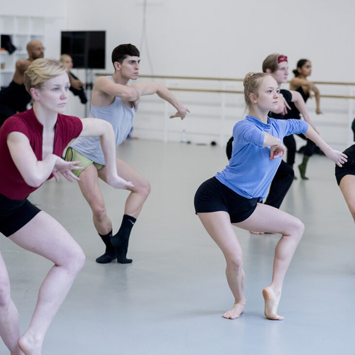 A group of dancers is practicing in a studio, performing synchronized movements with focused expressions. They are dressed in various workout clothes and appear to be concentrating on their routine.