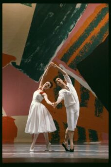 Two ballet dancers in white costumes perform a duet, holding hands and raising their arms. A colorful abstract backdrop is in the background.