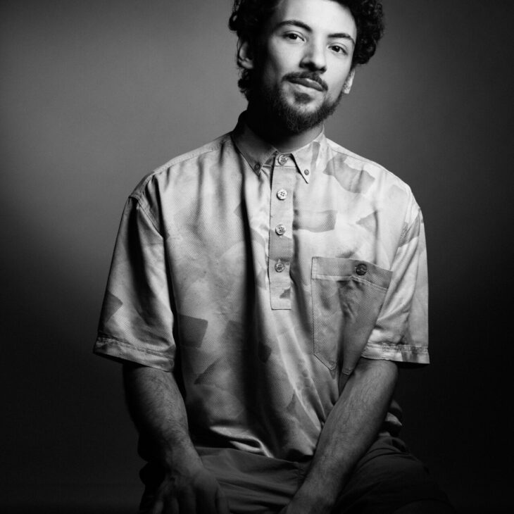 A man with curly hair and a beard is sitting and facing the camera, wearing a short-sleeve button-up shirt in a black-and-white photo.