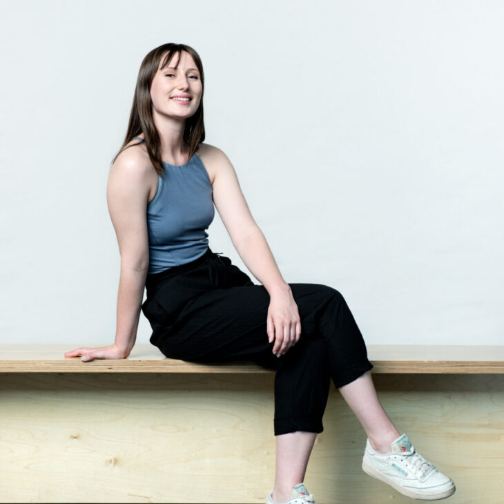 A person with shoulder-length brown hair sits on a wooden bench, wearing a blue sleeveless top, black pants, and white sneakers.