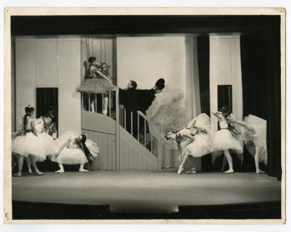 Black and white photo of dancers in ballet costumes on stage, with some dancing on the ground and others on a staircase.