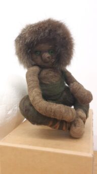 A handmade felted doll with a fur hood sits on a wooden box against a plain background.