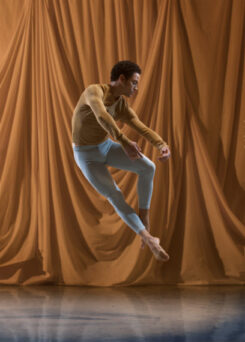 A person in a tan long-sleeve shirt and light blue pants performs a mid-air dance leap in front of a draped curtain backdrop.
