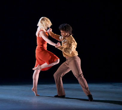 A woman in a red dress and a man in a beige jacket and brown pants are dancing together on a dimly lit stage.