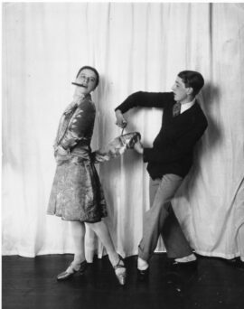 A man in a suit and tie mimics cutting a woman's jacket with scissors while she looks away, holding a cigar in her mouth. They stand against a plain white curtain backdrop.