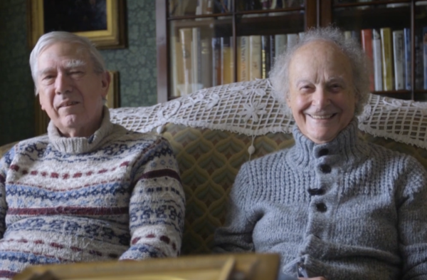 Two elderly individuals sit closely on a couch in a cozy room. They wear knitted sweaters and smile warmly at the camera, with a bookshelf in the background.